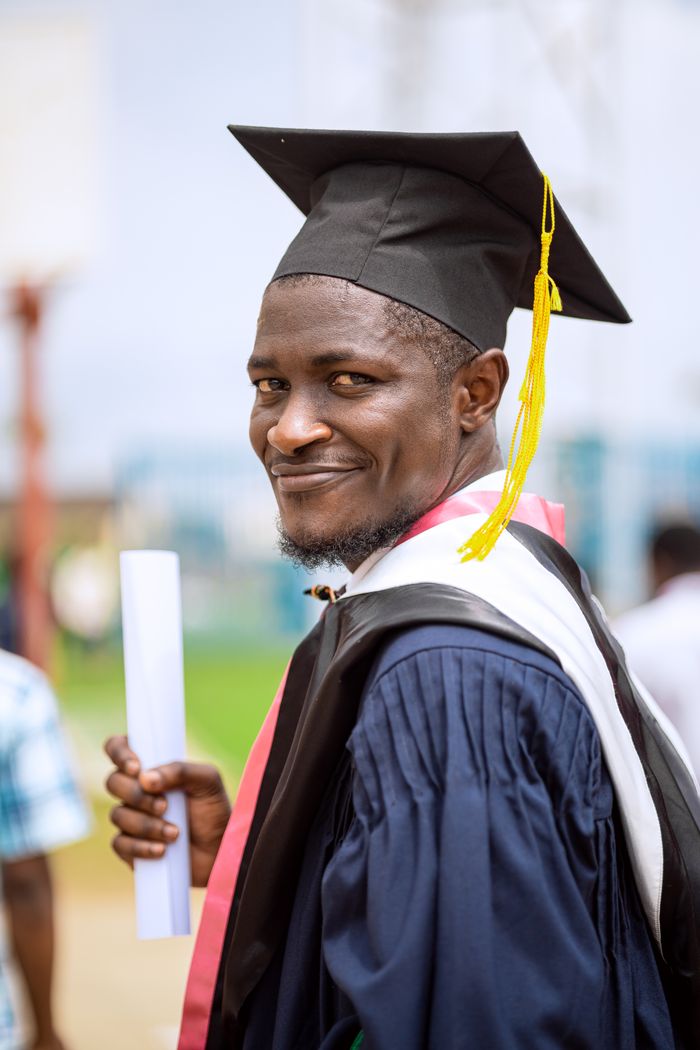 Proud graduate holding a diploma, smiling in a cap and gown during a university graduation ceremony outdoors.